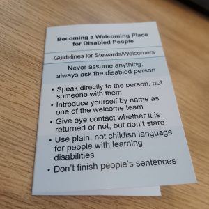 A Stewards Welcome Card with guidance for welcoming disabled people lies on a wooden table next to an open laptop.
