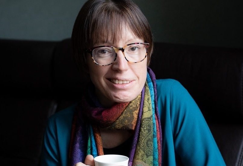 Smiling woman wearing glasses (Emily Owen) who is wearing a turquoise shirt and a colourful scarf, and holding a coffee cup