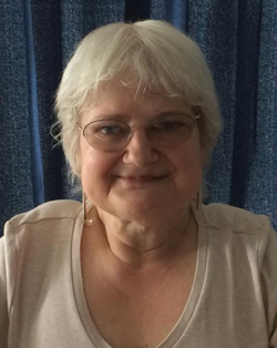An older woman (Jan Nicholson, Operations and Events Manager for TTR) with short white hair, glasses, and earrings smiles while sat in front of blue curtains.