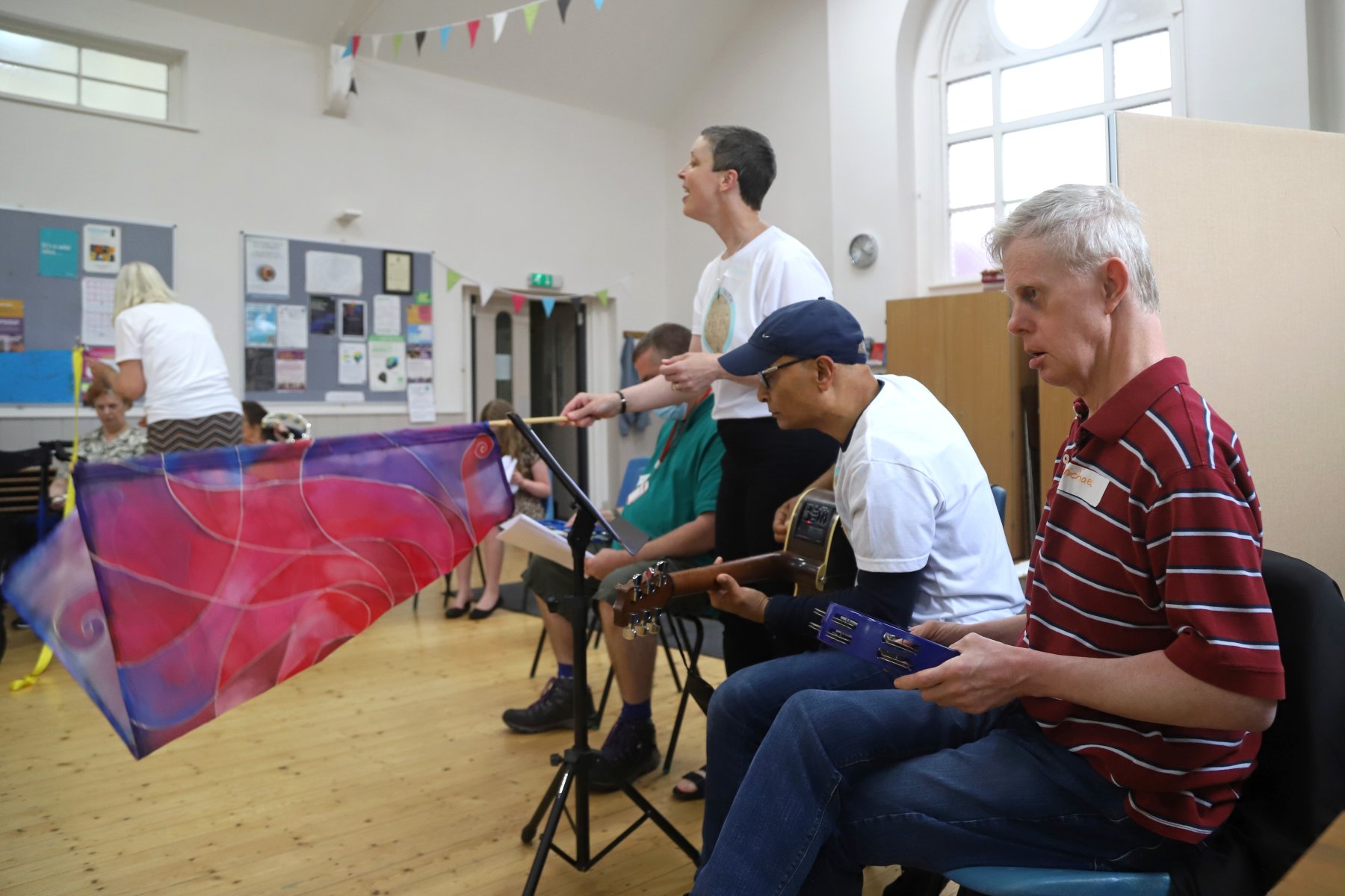 Adults play music and wave a colorful scarf together in a well-lit community room with wooden floors.