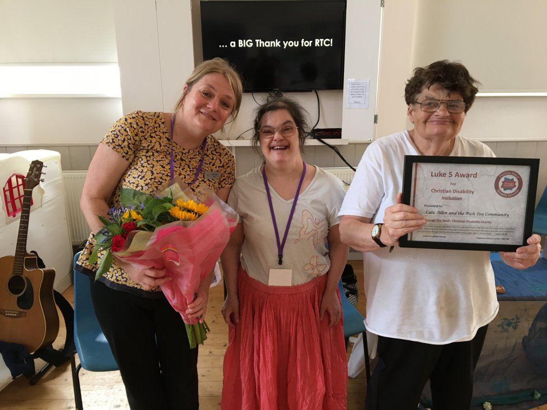 Three women (Cate, Sarah and Margaret) stand indoors smiling, with one holding flowers, another holding a Luke 5 Award certificate, and a TV in the background displaying the message Thank you for RTC!.