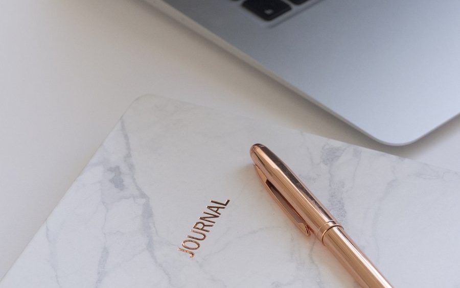 A marble-covered journal labeled JOURNAL sits on a white surface next to a gold pen and a silver laptop.