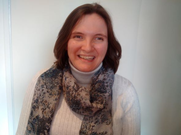 A woman (Katie Budd) with shoulder-length brown hair smiles while wearing a white sweater and a patterned scarf in front of a plain background.
