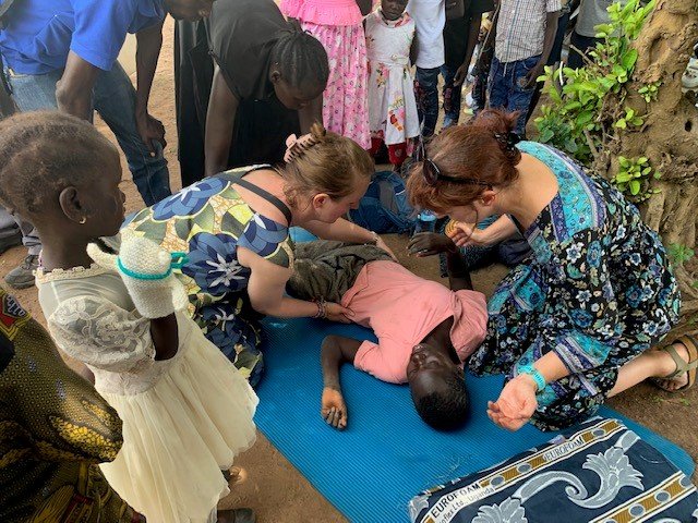 Two female UK therapists help a child who is lying on a mat outside while several people observe in the background.