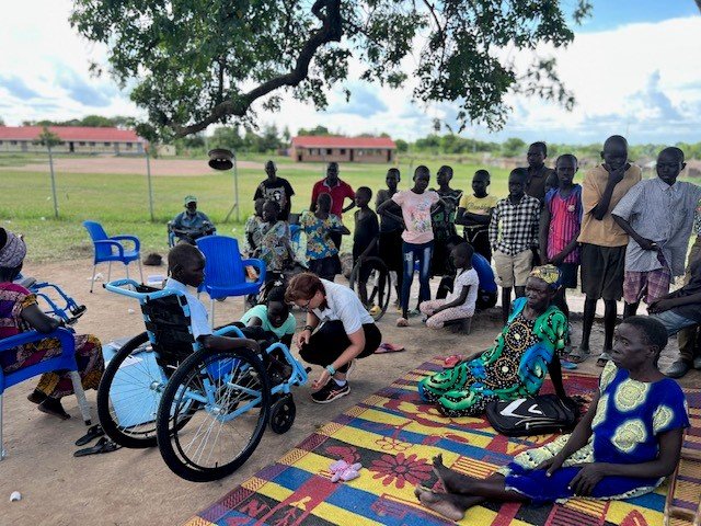 Outdoors, several people stand together as a woman assists a child in a wheelchair and others observe.