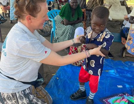 A female UK therapist assists a young Kenyan boy to stand up in new orthopedic boots, outside while several people watch in the background.