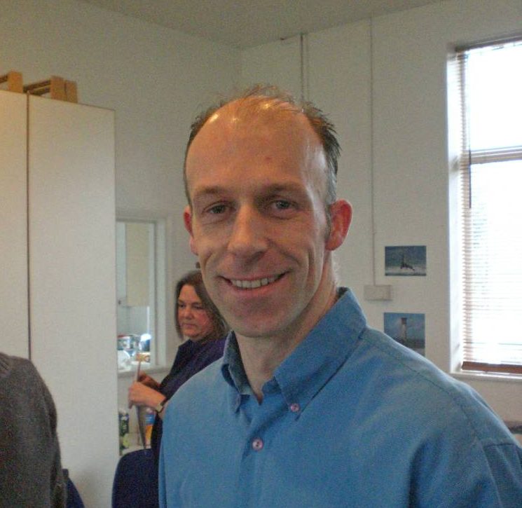 A man (Tim Wood, CEO of TTR) wearing a blue shirt and name tag smiles in an office while another person is visible in the background.