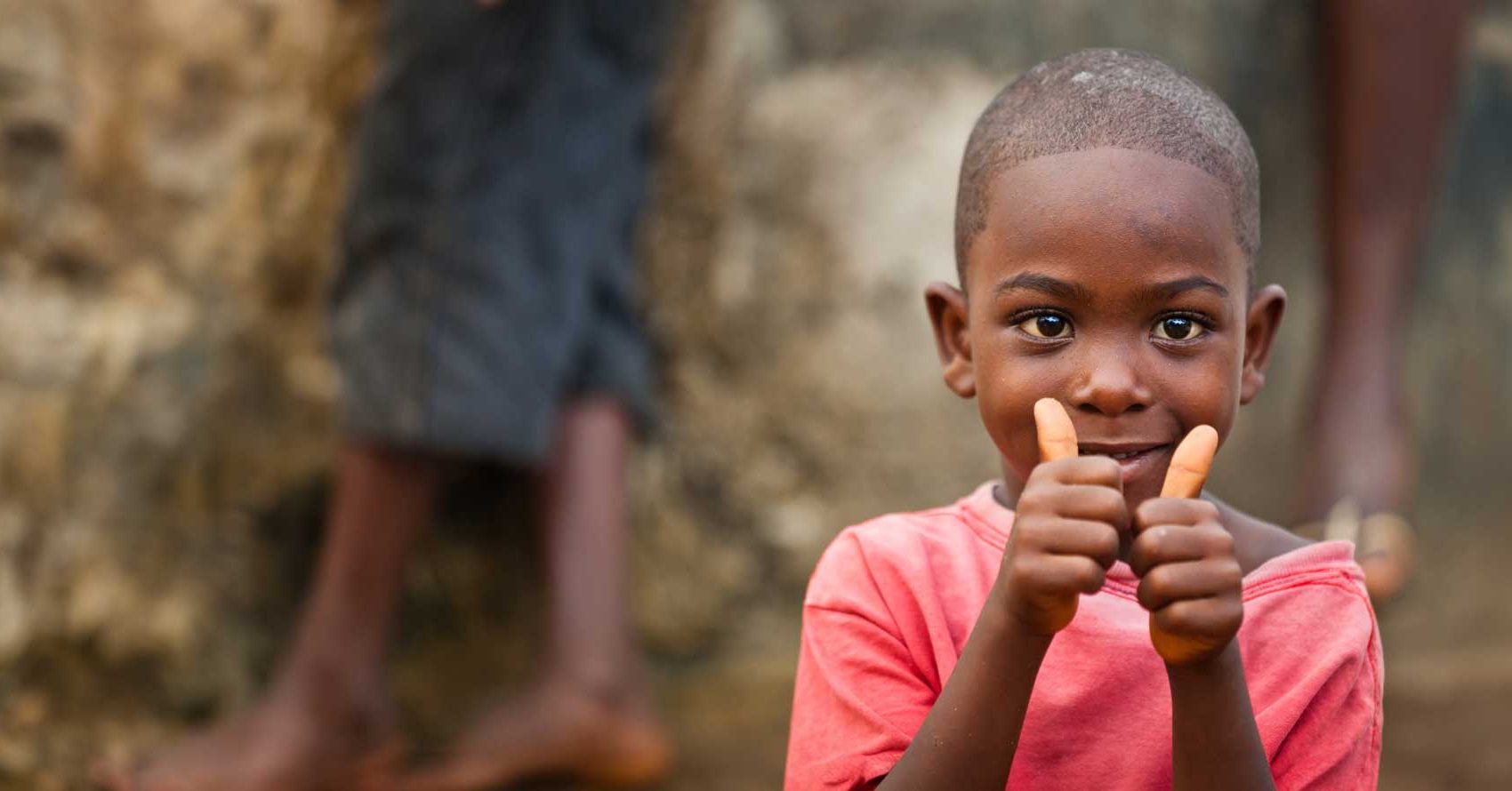 A young boy in a red shirt smiles and gives two thumbs up, standing in front of a blurred background with another person behind him.