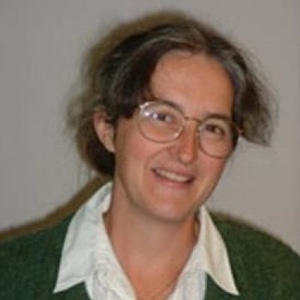 A woman wearing glasses with dark hair and a white shirt is smiling at the camera in front of a neutral background.