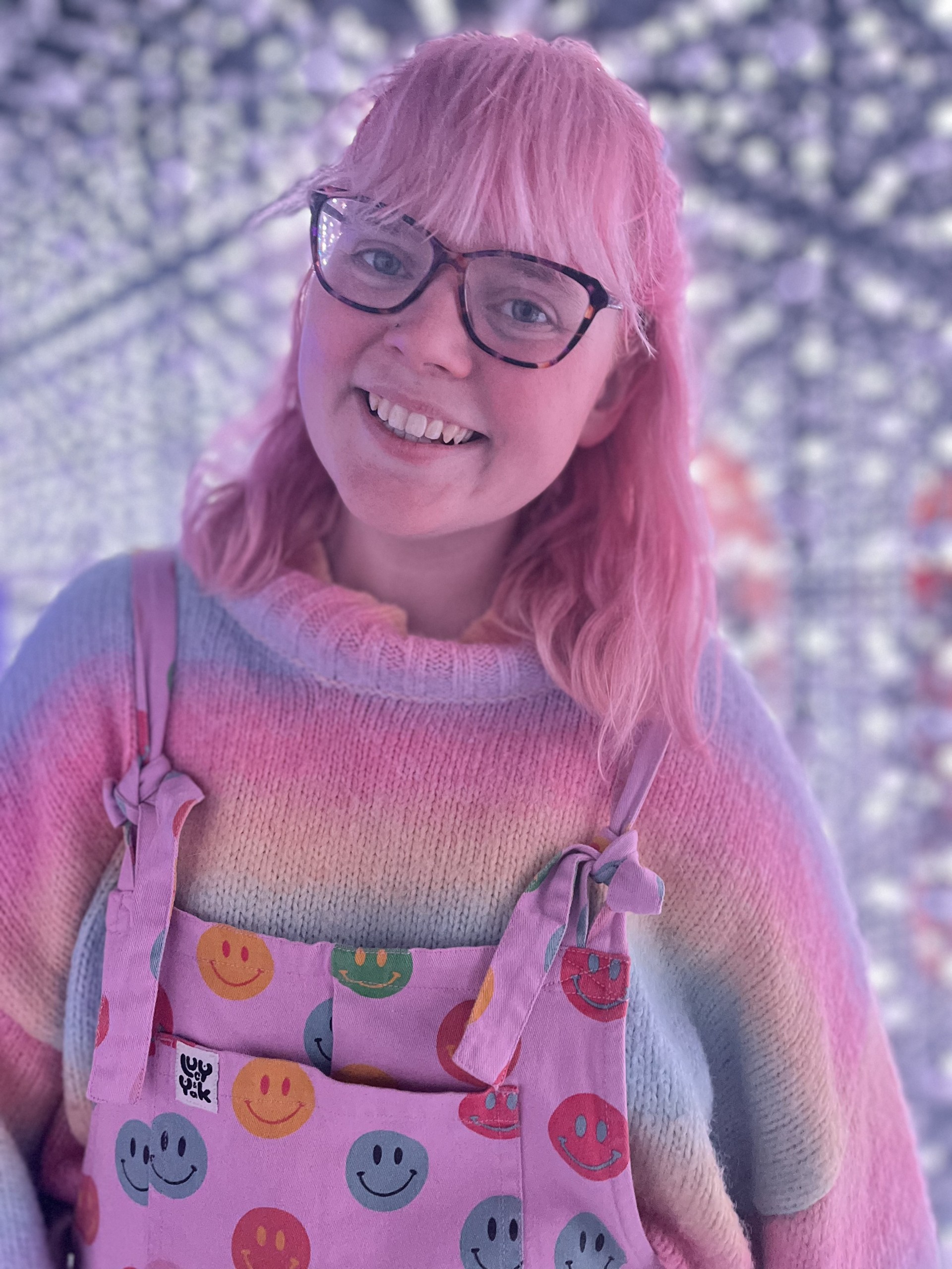 A person, Triona Brading, with pink hair and glasses smiles, dressed in colourful overalls with smiley faces and a rainbow-striped sweater.