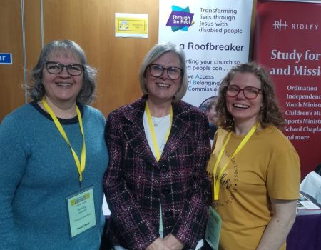 Three women with name badges smile and stand together in front of a banner that reads Be a Roofbreaker at an indoor event.
