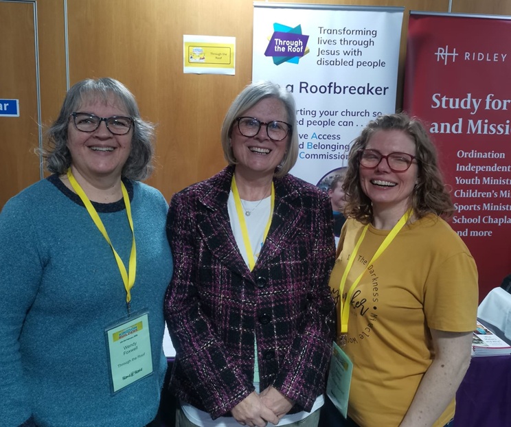 Three women with name badges smile and stand together in front of a banner that reads Be a Roofbreaker at an indoor event.