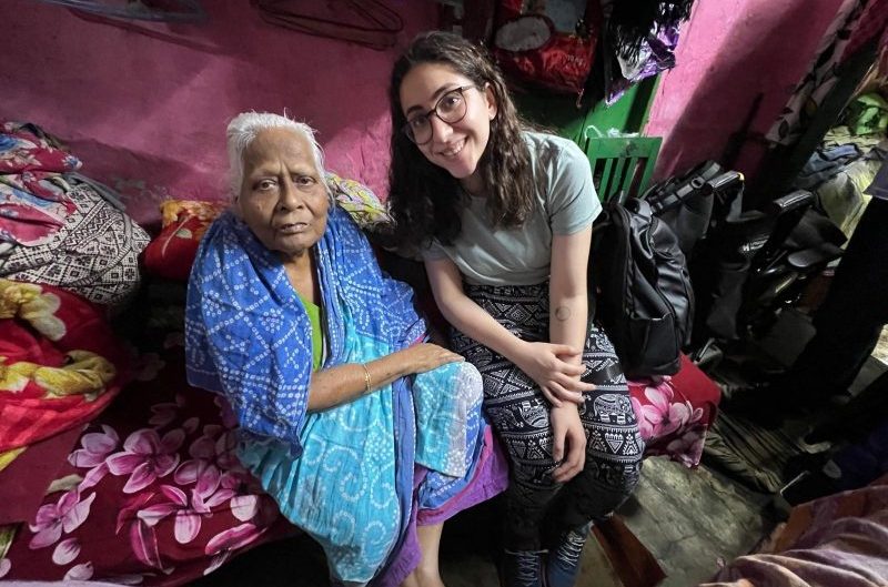 A young UK woman and an elderly Indian woman sit side by side on a bed in a colorful room, both smiling at the camera.