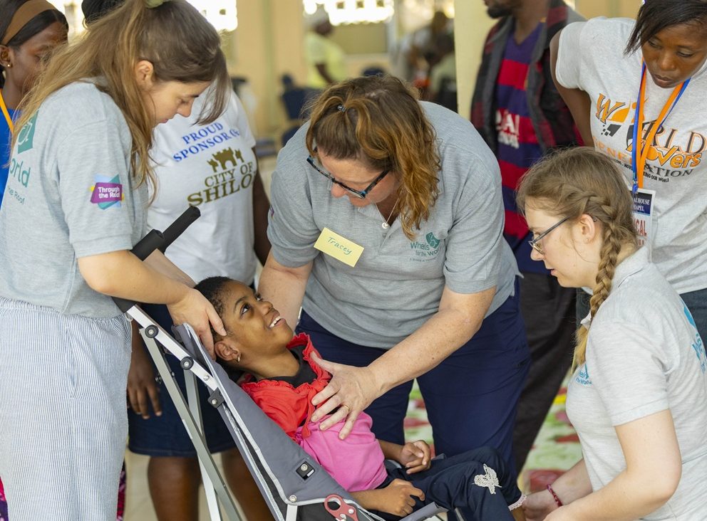 Several female therapists interact affectionately with a smiling Ghanaian child in a wheelchair, sat indoors. They are supporting him to be comfortable in the new chair.