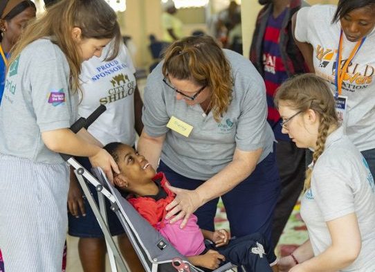 Several female therapists interact affectionately with a smiling Ghanaian child in a wheelchair, sat indoors. They are supporting him to be comfortable in the new chair.