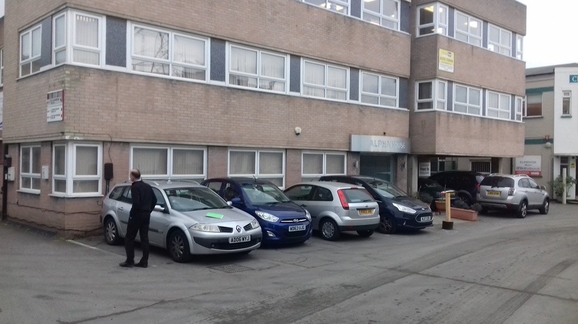 A man (Tim Wood, TTR CEO) is walking by parked cars in front of a beige office building (Alpha House in Morden) that has large windows.