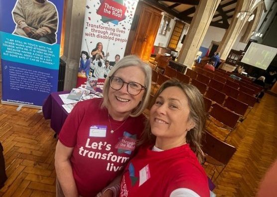 Two women wearing red shirts stand together and smile at a community event in a hall, with banners promoting church disability inclusion visible in the background.