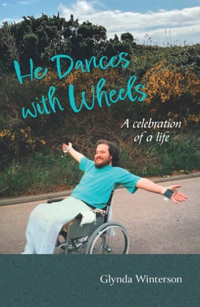 Book cover of 'He Dances With Wheels', showing a smiling man (David Winterson) in a wheelchair, stretching his arms wide and smiling, on a road by a hedge.