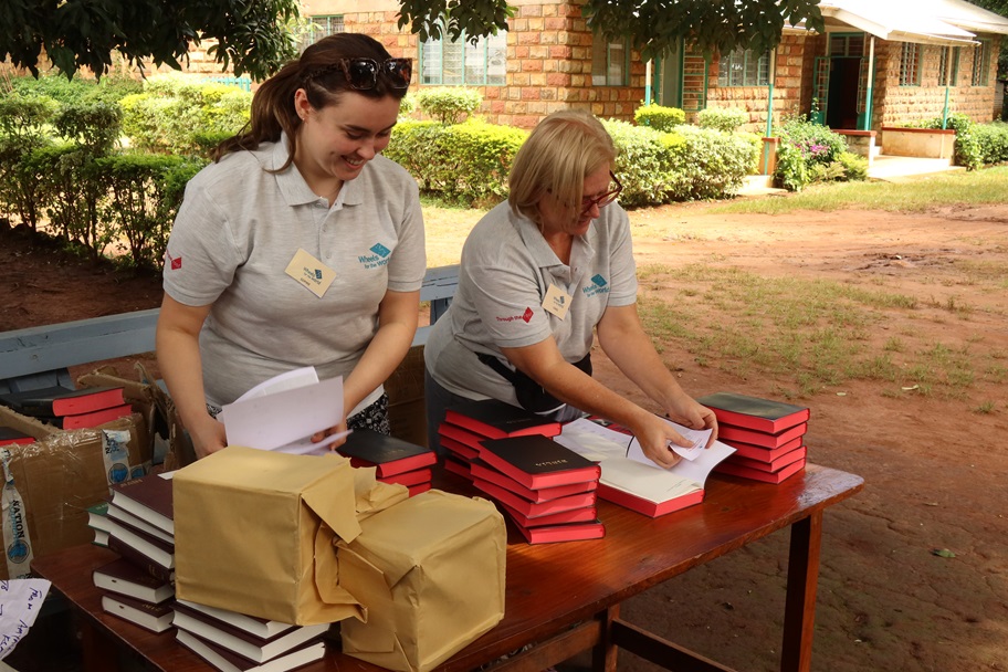 Two women wearing grey Wheels for the World polo shirts organize and stack bibles on a wooden table set outdoors by a garden and a building.