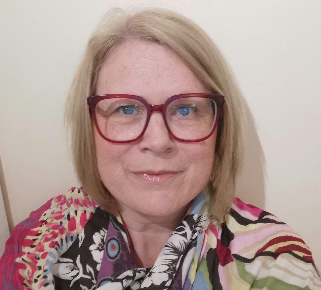 A smiling woman (Carol Dyer) with fair hair, wearing glasses and a colourful shirt, in front of a plain light wall.