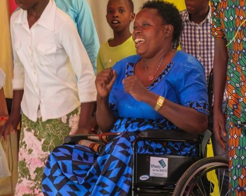 A woman in a blue dress sits smiling in a wheelchair among cheerful, standing people.
