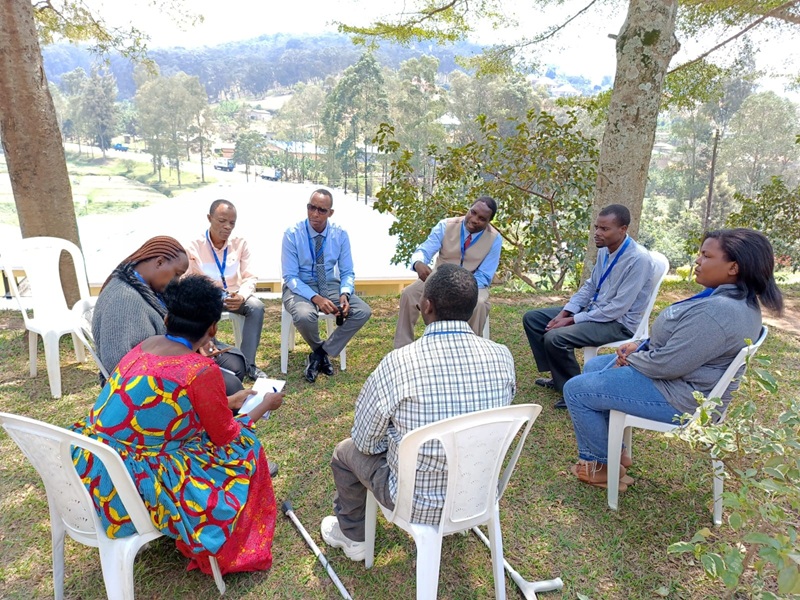 Eight Rwandan people are seated in a circle on white chairs outside, conversing beneath trees.