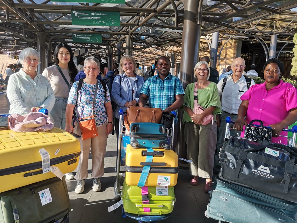 Eight people stand smiling at an airport with their luggage and suitcases under a glass roof, preparing for their Wheels for the World journey.