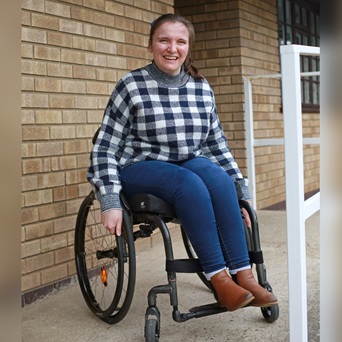 A woman in a wheelchair smiles while wearing a checkered sweater, jeans, and brown boots next to a brick building.