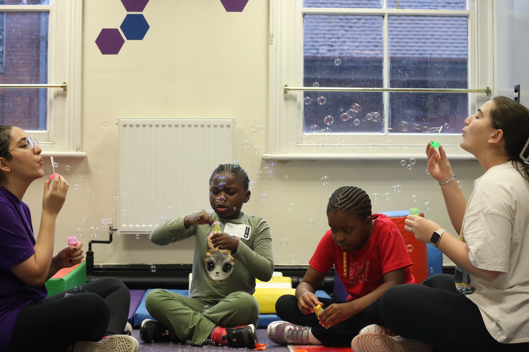 Two young adults and two children sit on the floor in a church hall, blowing bubbles and playing together.