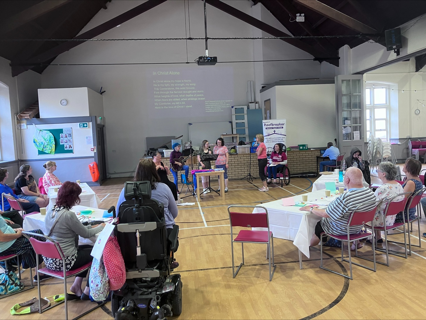 About 15 people seated at tables in a church hall listen to speakers at the front near a screen and projector during a Roofbreakers initiative event.