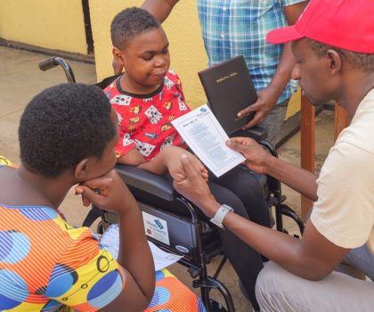 A man in a red cap prays for a young Rwandan girl in a wheelchair while a woman looks on and another man holds a leather bound Bible.