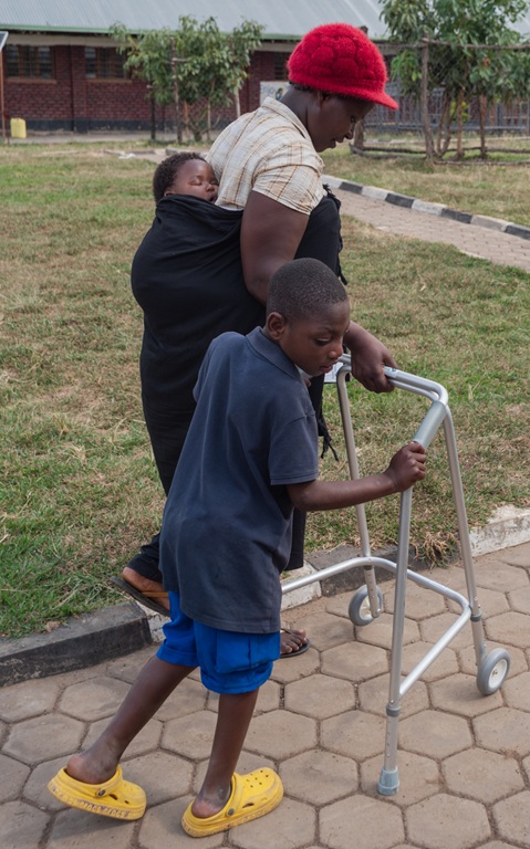 A Rwandan woman carries a sleeping baby while a boy uses a metal walker during Wheels for the World Rwanda 2026.