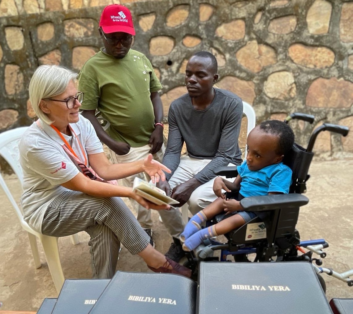 A UK female Wheels for the Word volunteer shows a book to a young Rwandan boy in a wheelchair, joined by two men at the Wheels for the World Rwanda 2026 distribution.