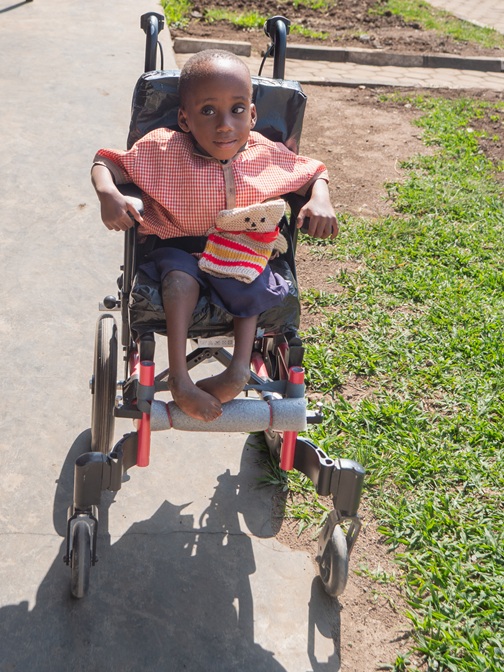 A young Rwandan boy, Pabrico, sits in a Wheels for the World Rwanda 2026 wheelchair while holding a small knitted teddy bear.