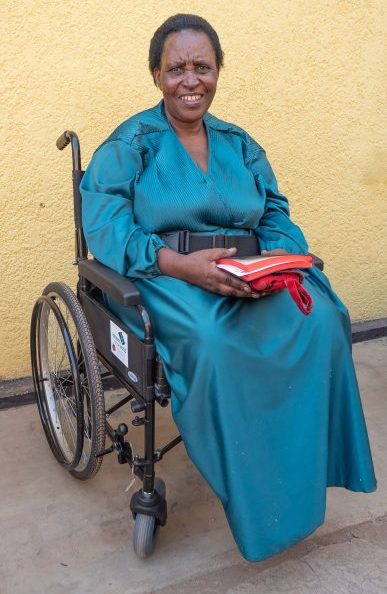 A smiling Rwandan woman in a teal dress sits in a wheelchair holding a Bible.