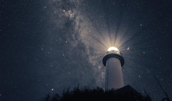 A lighthouse emits beams of light at night beneath a sky full of stars.