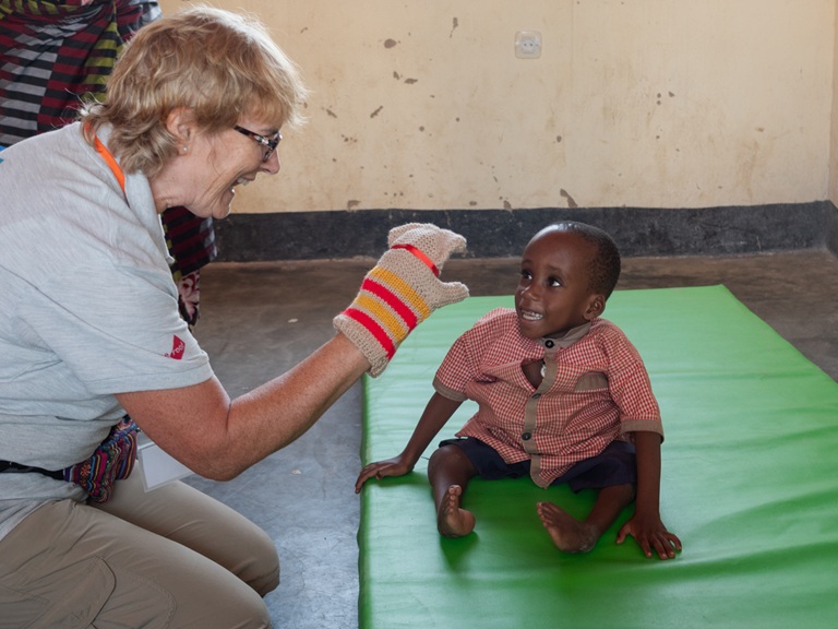 A female UK Wheels for the World volunteer (Moira) plays with a sock puppet for a smiling Rwandan boy, sat on a green mat during the Wheels for the World Rwanda 2026 mission.