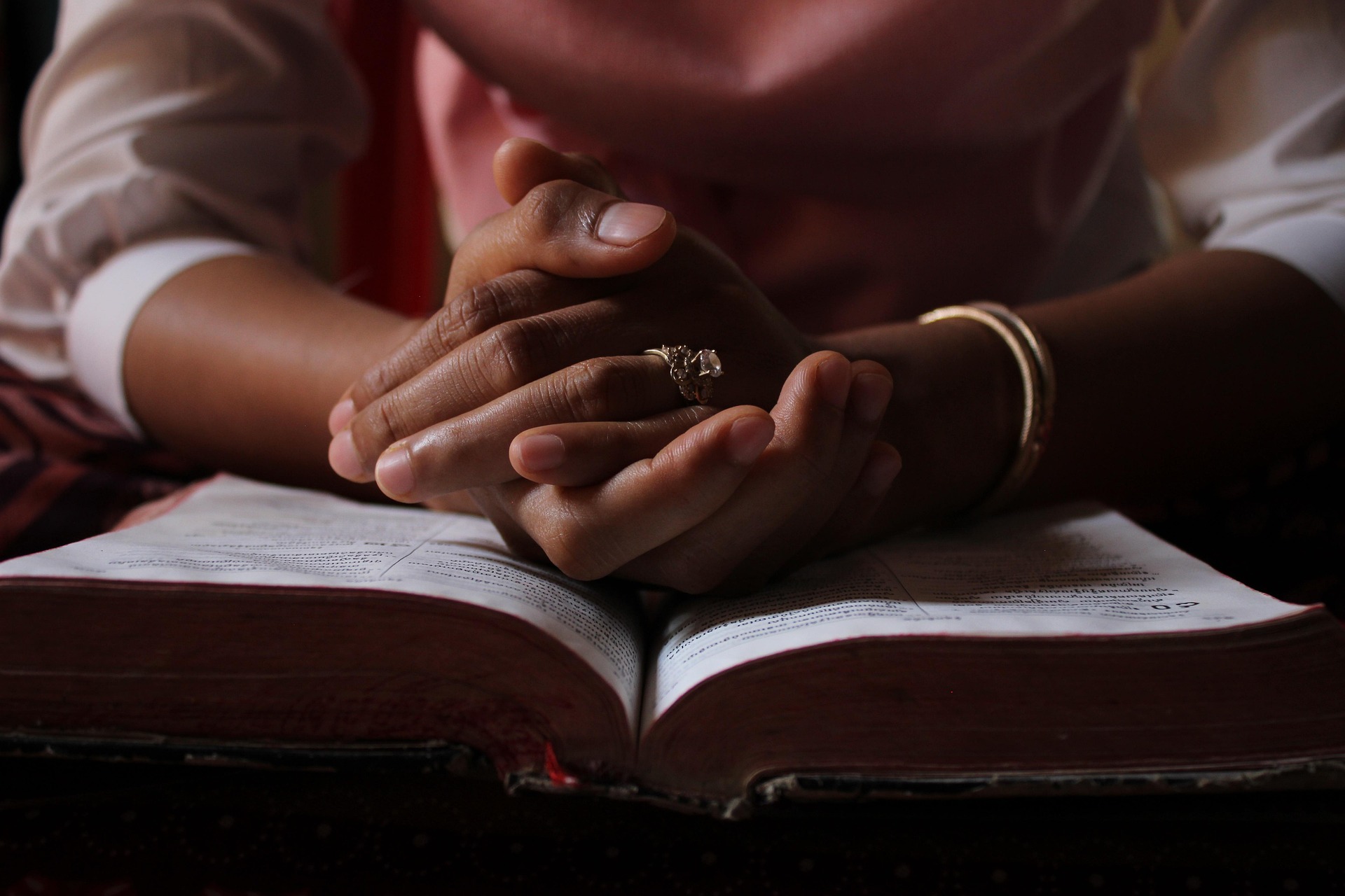 Hands wearing a ring are clasped in prayer over an open book that may be a Bible, softly illuminated.