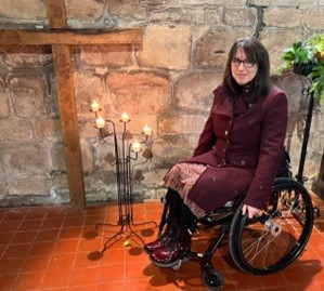 Lynda sits in her wheelchair in front of a stone church wall. To her left, there's a large wooden cross and a few candles in a floorstanding candle holder.