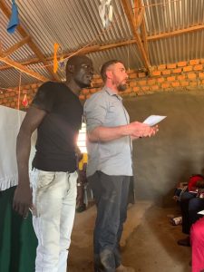 David, a member of the Wheels for the World team, preaches while holding his notes. A colleague from Uganda stands next to him, helping and translating.