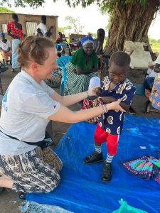 A member of the wheels team helps Peter, a five year old boy, to stand up in his new special boots. He's smiling at the camera, while his mother looks and smiles at him from behind.