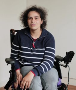 Photo of Ashleigh with black curly hair, sat in her wheelchair, wearing a blue and white stripey jumper.