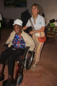 A smiling UK team-member is shown pushing the newly received wheelchair of a Kenyan lady who has a big broad smile on her face.