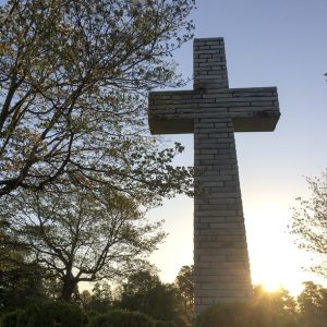 Photo of a Christian cross built from bricks, standing in a garden with trees and bushes around it, and the sun rising behind it