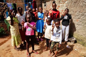 A big group of Kenyan children waving at the camera, stood in an outdoor yard, with washing hanging behind, next to a Kenyan lady in a Sunday-best yellow dress