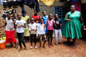 A big group of Kenyan children singing and dancing in an outdoor yard, with washing hanging above them, led by a Kenyan lady in her Sunday-best green dress