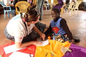 A female UK Wheels team member is sitting on the floor with a smiling young Kenyan boy. They are talking and using paper from a notebook. There are coloured crayons and coloured fabric on the floor– perhaps a play parachute, with stripes of red, yellow and purple. 