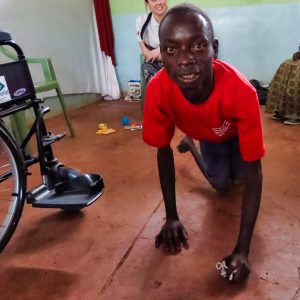 Photo of Sammy, a young man with cerebral palsy, wearing a red T shirt. He is crawling on his knees, next to his new wheelchair, given to him on the Wheels for the World Kimilili mission to Kenya. Behind him a female UK Wheels team member is chatting to a Kenyan lady.