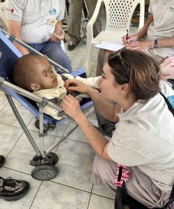 A female UK Wheels volunteer (Sophie the play worker?) is sat at eye level with a disabled Kenyan child who sat in a push-chair. The child has an enlarged head and a very small torso with little or no limbs. She is wrapped is a soft cream blanket, and the Wheels worker is clipping her lovingly into the push-chair to keep her secure with the safety belt.