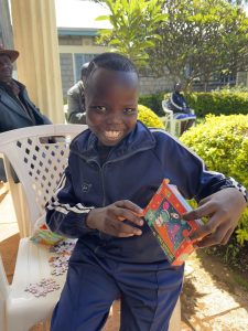 A young Kenyan girl in a dark blue tracksuit is smiling at the camera, holding the box of a space jigsaw puzzle she’s been given to play with. 